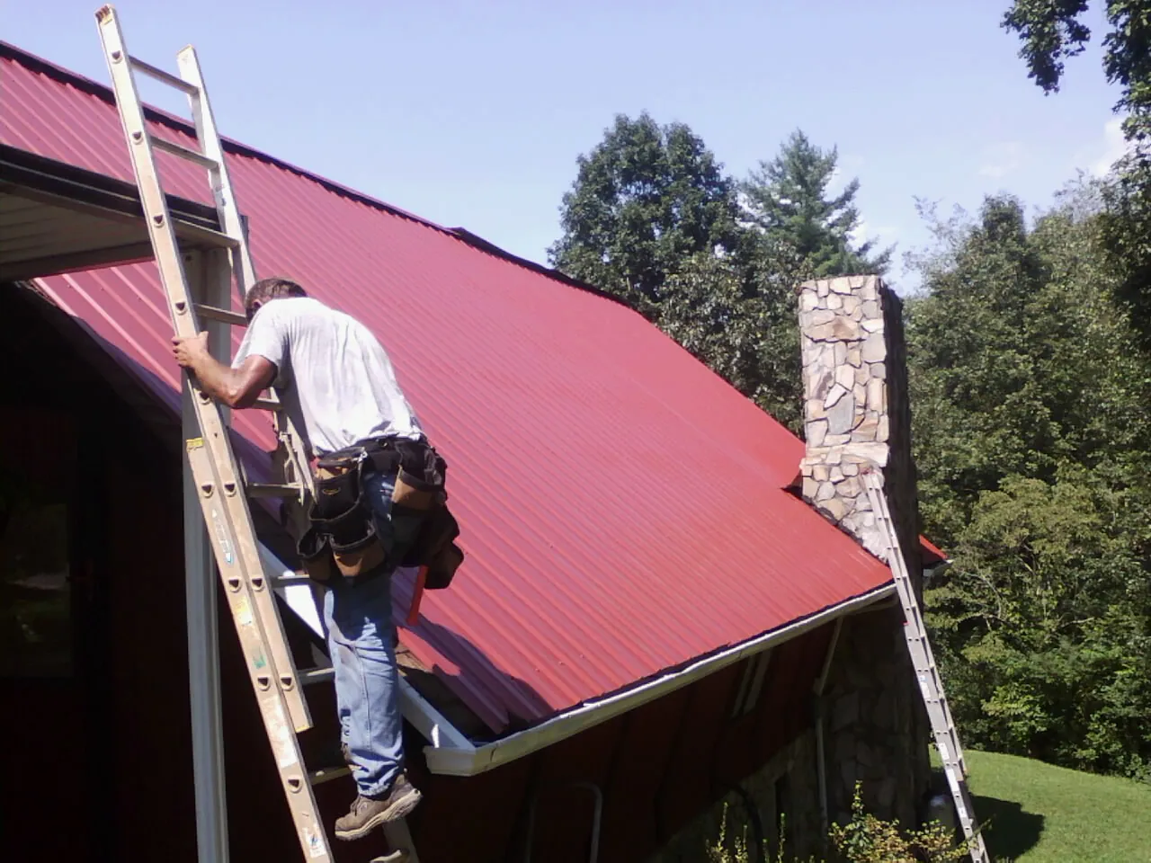 Roofer on a ladder beside a red metal roof and stone chimney — Soffit & Fascia Repair by Roofing Brothers in Villa Ridge